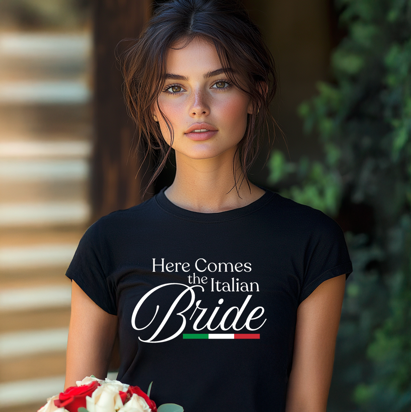 Woman wearing a black t-shirt with 'Here Comes the Italian Bride' text, holding flowers. From a matching bridal party t-shirt collection for the whole family.