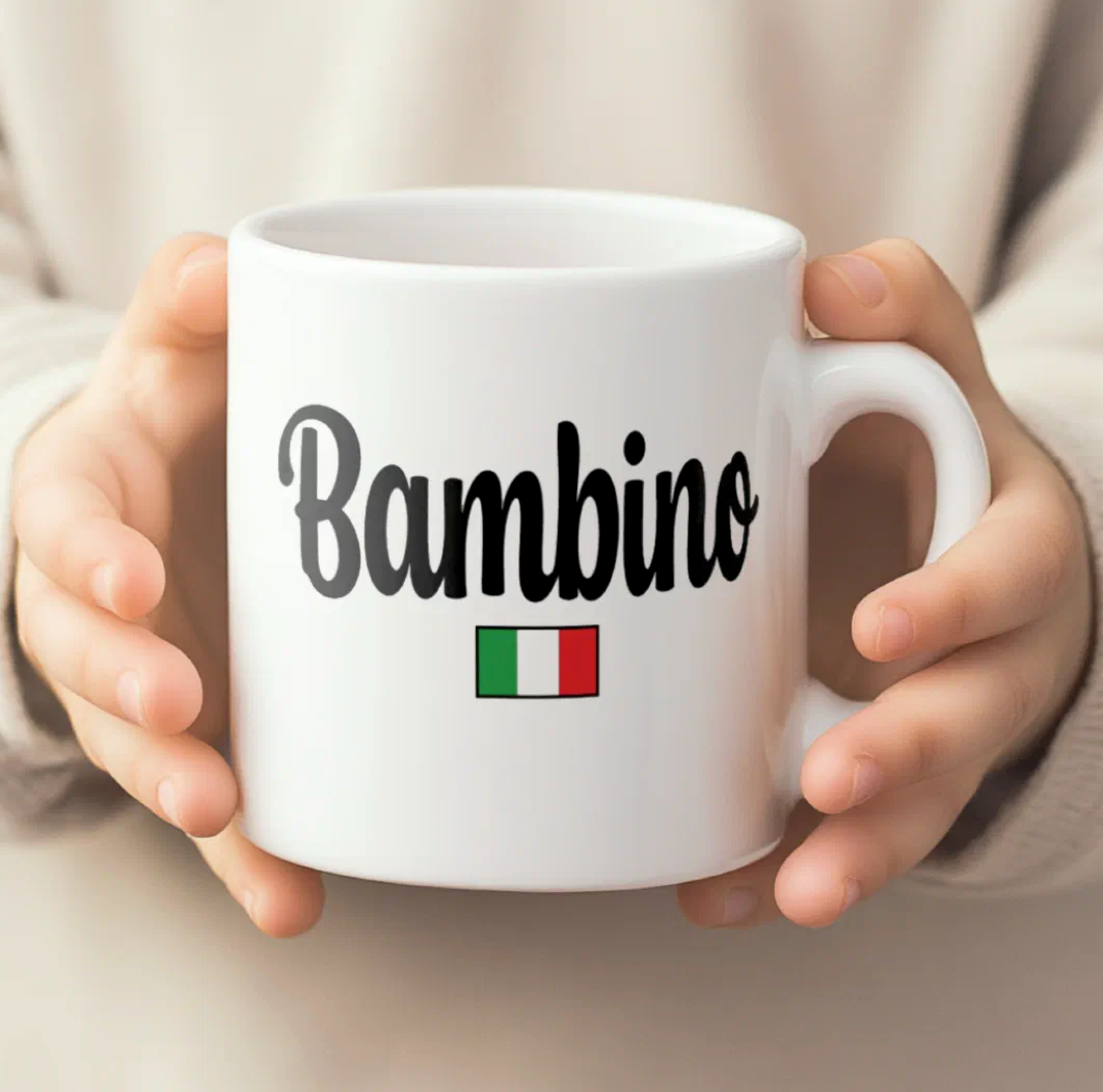 White mug with 'Bambino' and Italian flag held by hands against a neutral background