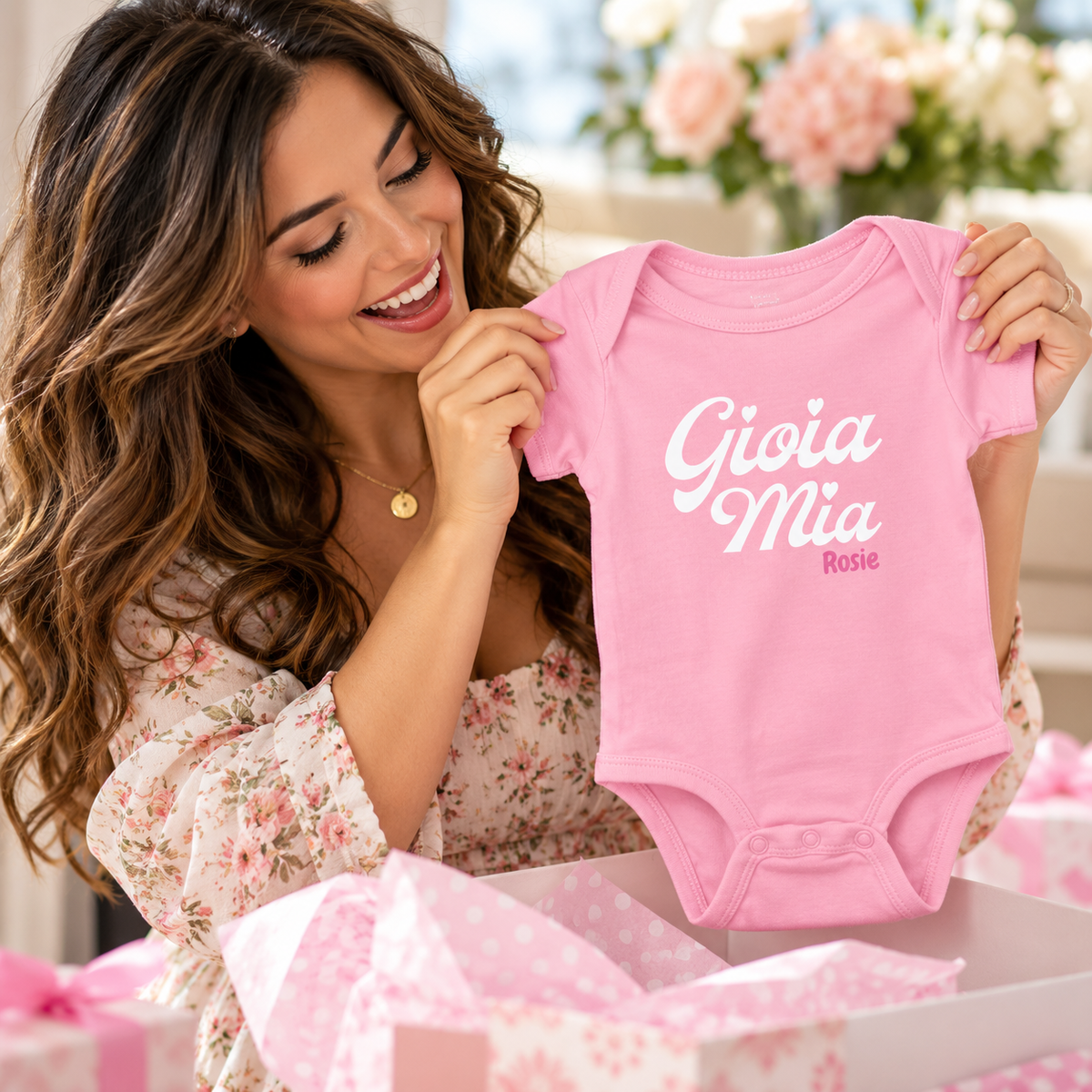 Happy expectant mom holding a personalized pink Gioia Mia Rosie Italian baby bodysuit at a baby shower.
