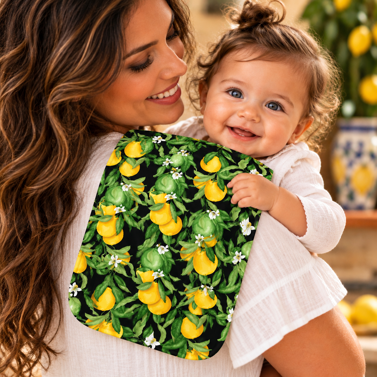 Smiling mother holding her happy baby over her shoulder with a Sicilian lemon-print burp cloth featuring yellow lemons, green leaves, and white blossoms on black.