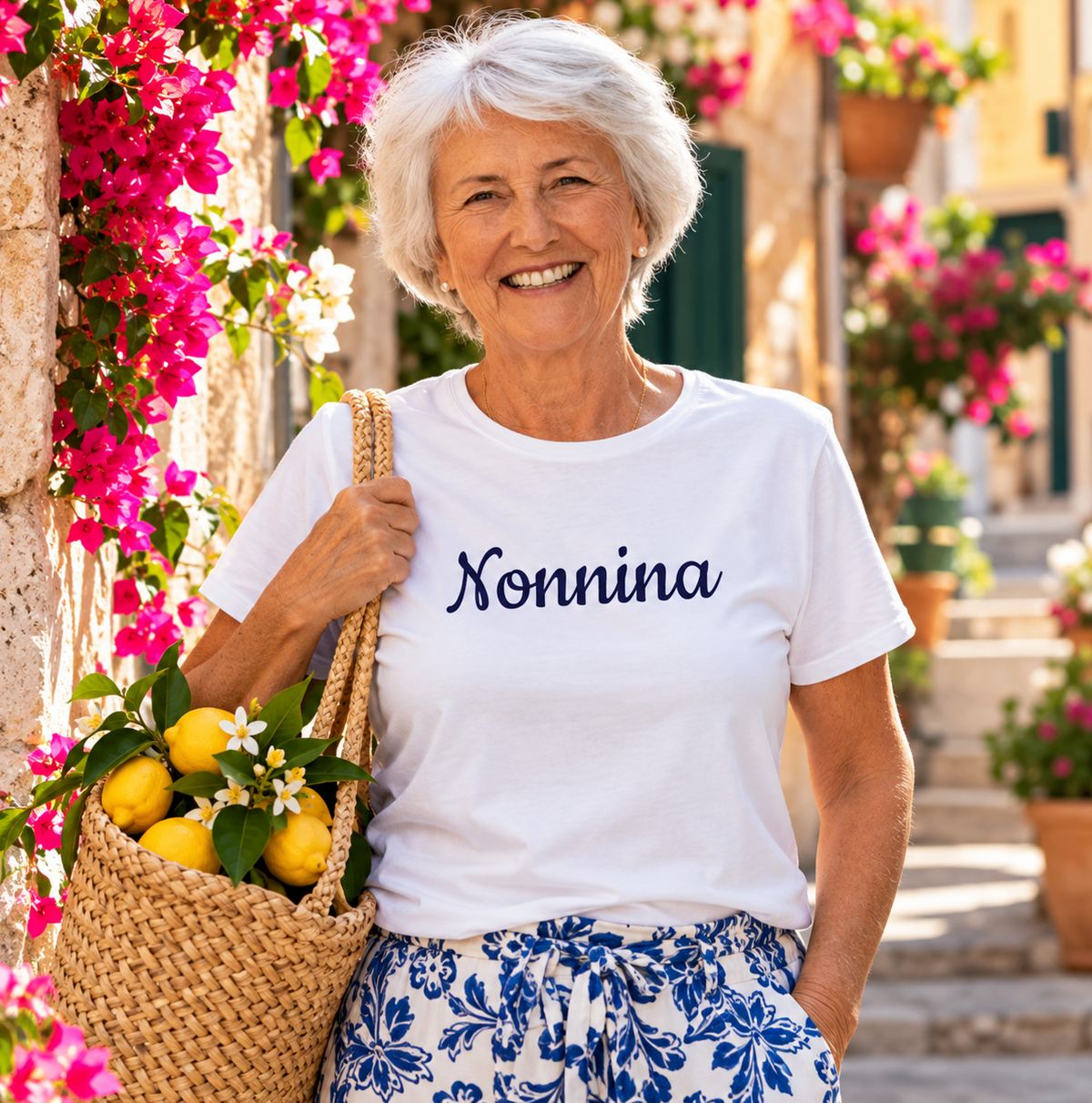 Smiling Italian grandmother wearing a white “Nonnina” T-shirt in a sunny Mediterranean village, carrying a woven basket filled with fresh lemons and surrounded by colorful bougainvillea.