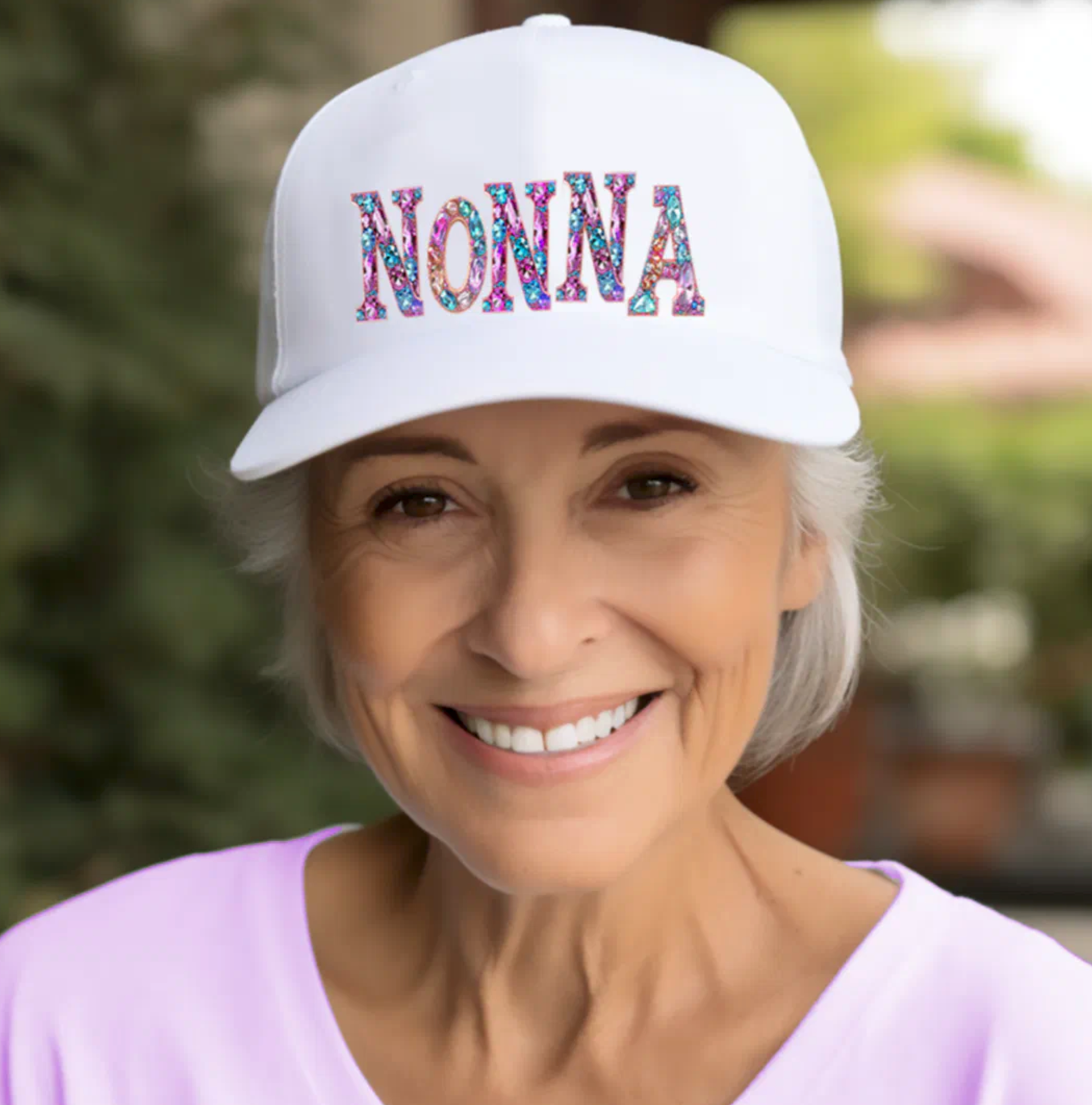 Woman wearing a white cap with 'NONNA' faux jewels in colorful letters, outdoors.