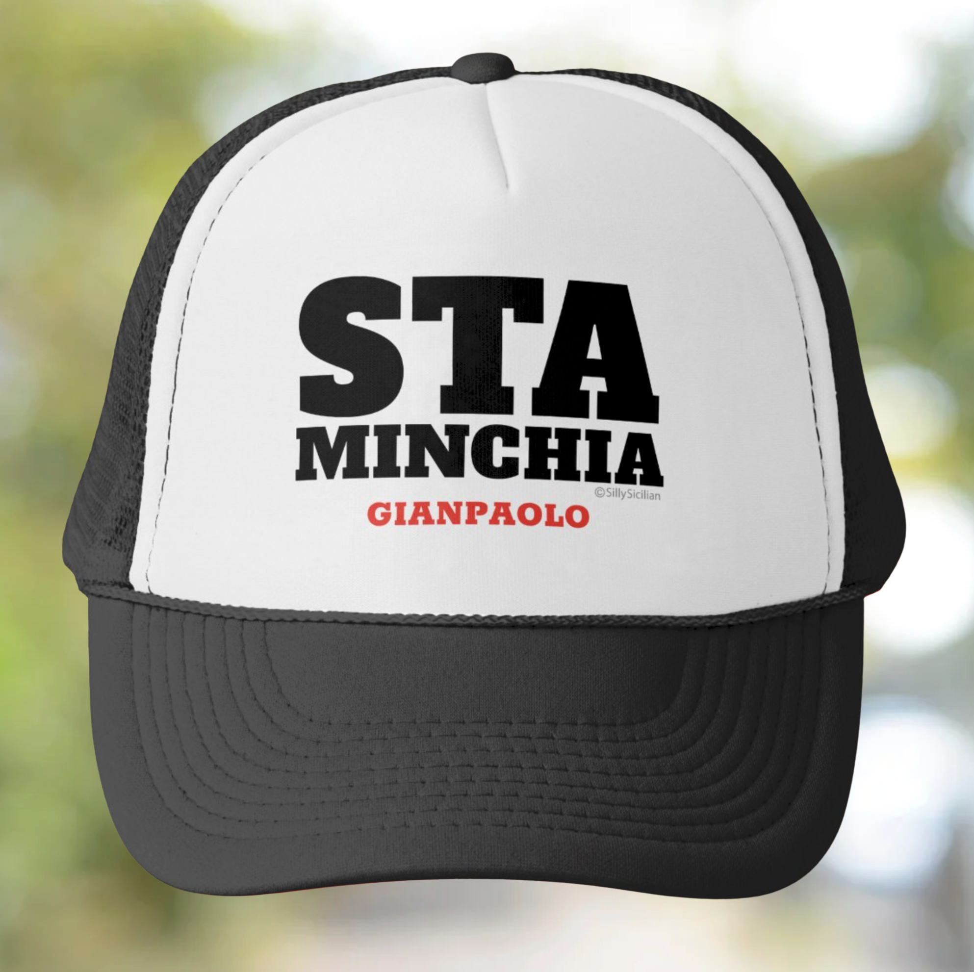 Black and white cap with Sta minchia personalized text on a blurred natural background