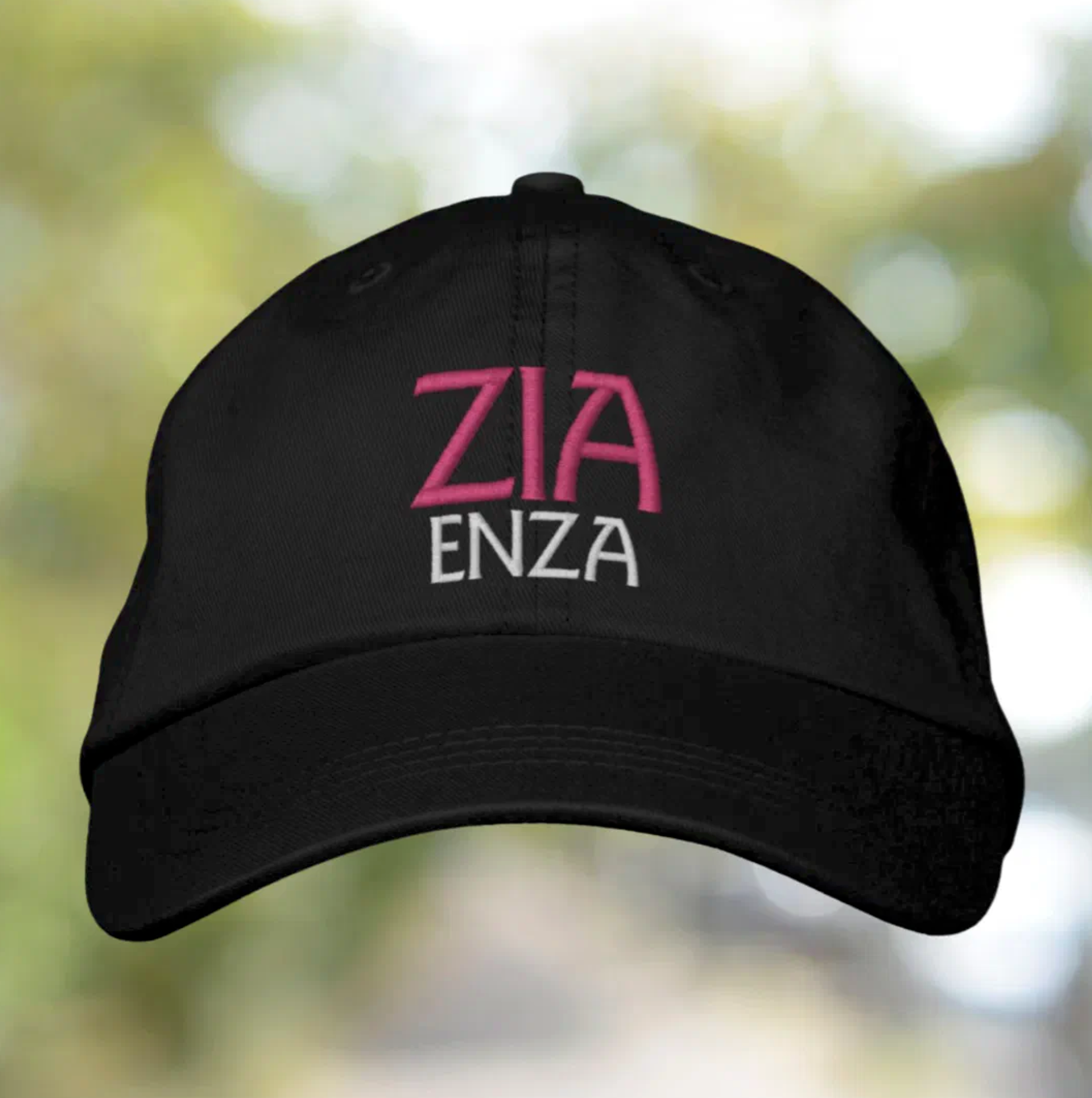 Black cap with 'ZIA ENZA' text in pink and white on a blurred natural background