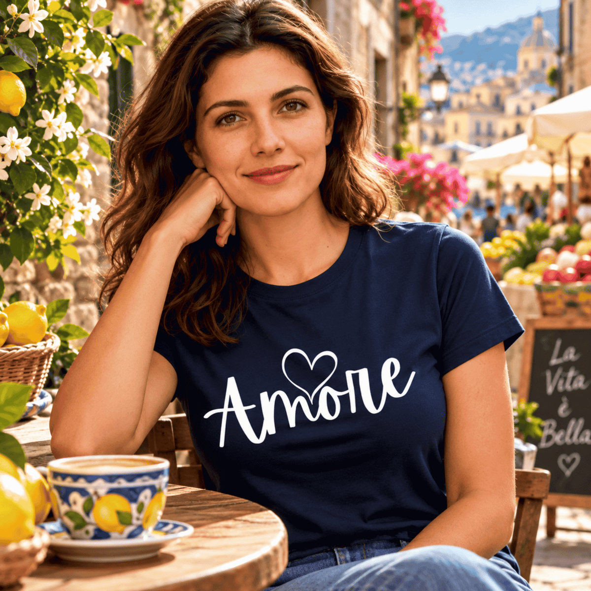 Woman wearing a navy blue Amore T-shirt with white heart lettering, enjoying cappuccino at a charming Italian village café surrounded by lemons and flowers. A romantic Italian-inspired gift for women, Valentine's Day, anniversaries, or lovers of Italian culture.