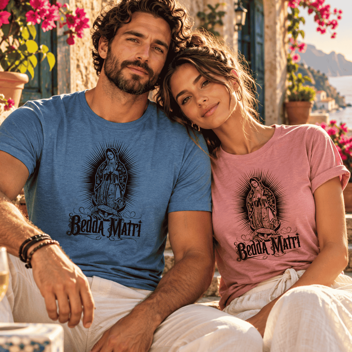 Couple wearing matching Bedda Matri Sicilian T-shirts in heather blue and dusty pink, featuring a vintage Madonna design, in a romantic Italian coastal setting.