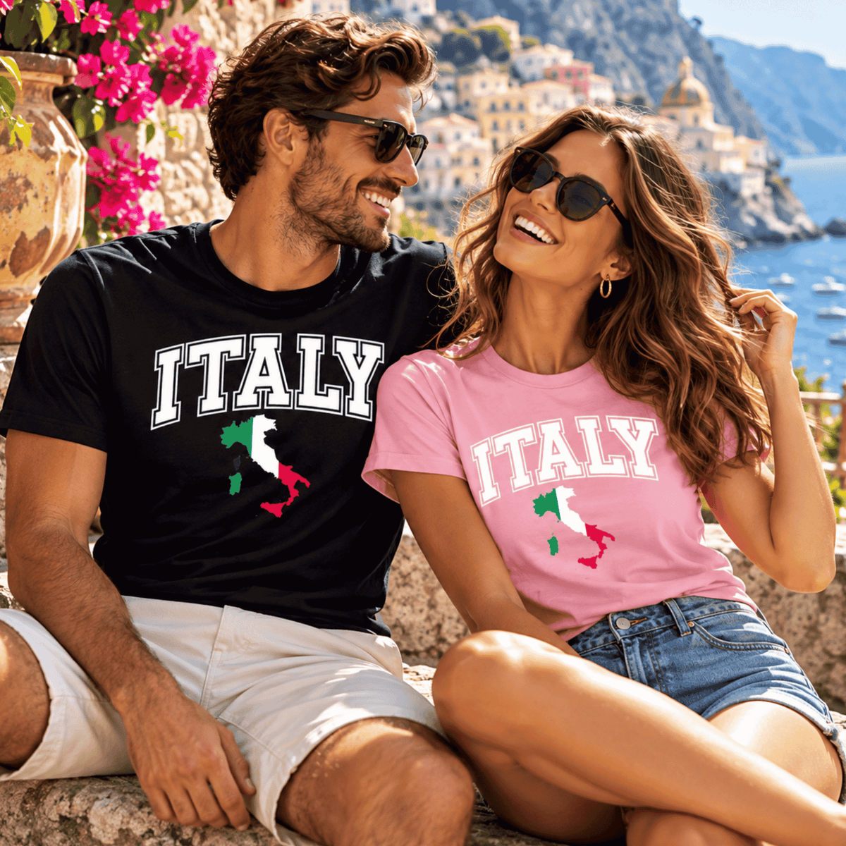 Couple wearing matching Italy T-shirts in black and pink, featuring the Italian flag map design, relaxing on a sunny Mediterranean coastal terrace.