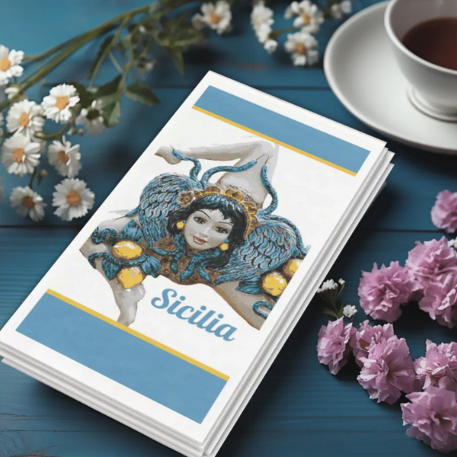 Stack of cards with a decorative design and 'Sicilia' text, surrounded by flowers and a cup of tea on a blue surface.